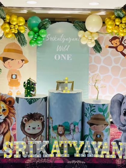 A safari-themed "Wild One" party featuring a little explorer character and friendly jungle animals. The setup includes an arched backdrop, custom-printed plinths, and a full balloon garland in green and gold.