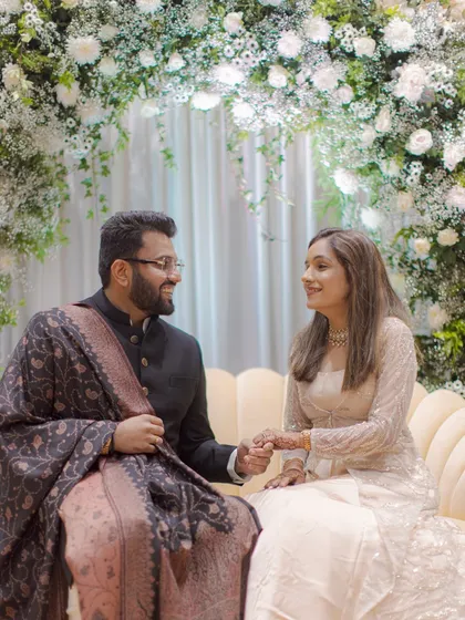 The couple shares a tender moment, seated on a couch under a beautiful white floral arch during their engagement ceremony.