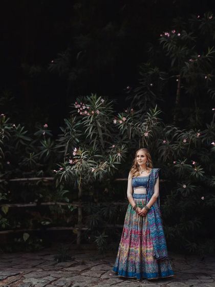 A stunning bridal portrait against a dark, moody backdrop of trees. The contrast makes the bride's colorful lehenga stand out beautifully.