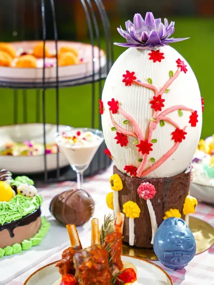 A large, beautifully decorated white chocolate Easter egg alongside other festive cakes and treats. The vibrant colors and detailed decorations create a magical and indulgent scene.