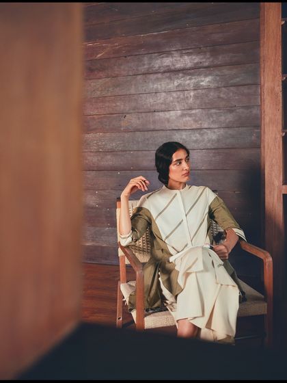 Seated in a wooden chair, the model's pose is relaxed yet elegant. The earthy tones of her outfit blend seamlessly with the wooden architecture and the green foliage outside.
