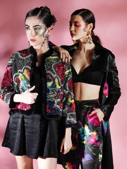 A dual-model editorial shot featuring heavily embellished and printed jackets. The styling is layered and complex, with artistic makeup creating a mystical, avant-garde aesthetic.