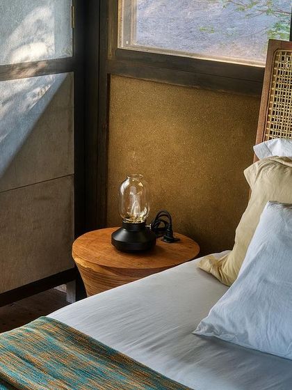 A bedside detail in the prefab cottage, where the texture of the earthen wall and cane headboard create a tactile and calming atmosphere.