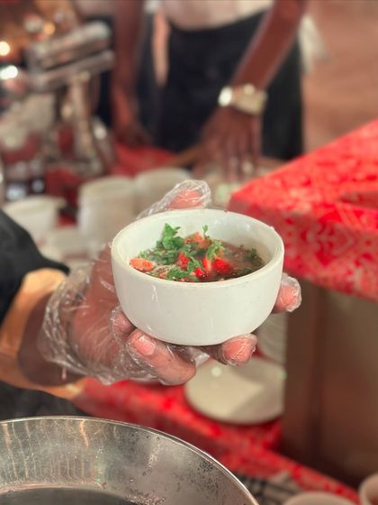 A close-up of a bowl of Thai soup being served. The clear broth is filled with fresh herbs and chilies, promising a spicy and aromatic experience.