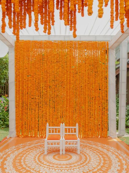 A stunning Haldi stage featuring a dense wall of bright orange marigolds. The custom-printed floor with a mandala design and traditional white chairs complete this vibrant and picturesque setup.