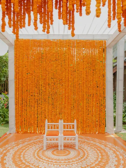 A stunning Haldi stage featuring a dense wall of bright orange marigolds. The custom-printed floor with a mandala design and traditional white chairs complete this vibrant and picturesque setup.