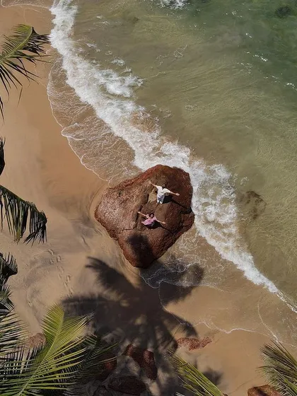 A beautiful drone view of a couple relaxing on a large rock by the sea, framed by palm trees. This kind of shot captures the tranquility and tropical beauty of a destination like Goa.