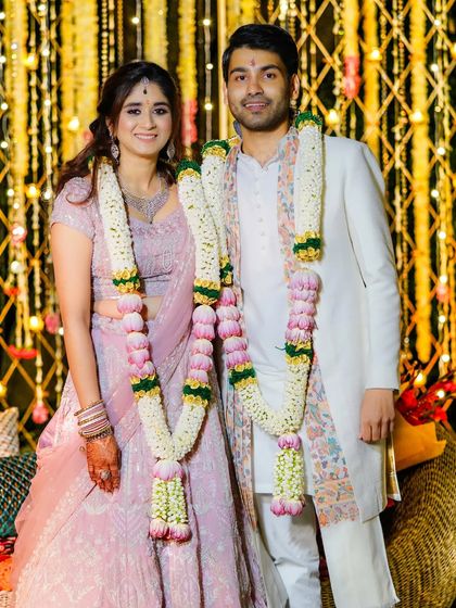 "Together is a wonderful place to be." The newly married couple, adorned with beautiful varmala garlands, posing for a classic wedding portrait against a backdrop of festive lights.