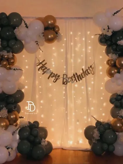 A simple and elegant backdrop for a home celebration. The setup features a balloon frame in dark green, white, and gold against a curtain of fairy lights.