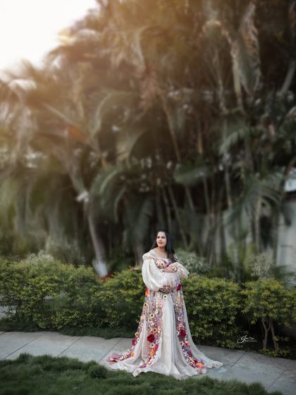 A wide shot showing the beautiful scale of the outdoor location. The mom-to-be stands peacefully in her floral gown, framed by tall palm trees and soft, natural light.
