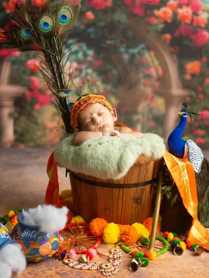 A divine Little Krishna asleep in a basket, surrounded by peacock feathers and marigolds. The garden backdrop adds a touch of Vrindavan's magic.