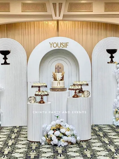 An elegant and serene stage for a First Holy Communion. This setup uses white 3D ridged panels, tall pedestals for the cake and offerings, and lush white and gold floral arrangements for a sophisticated look.