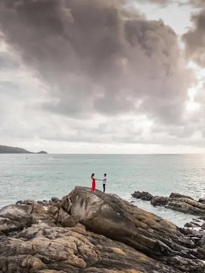 Harman and Komal stand together on the rocks, framed by a dramatic sky and the vast ocean. This is a perfect example of an epic destination portrait.