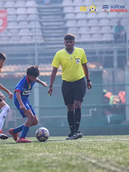 A young player demonstrates composure on the ball under the watchful eye of the referee during an AIFF Youth League match.