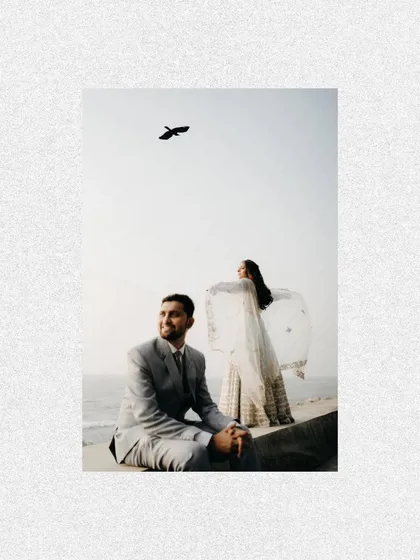 A creative shot from a beachside pre-wedding session. The groom sits in the foreground while the bride stands behind him with her dupatta flying in the wind, like a scene from a movie.