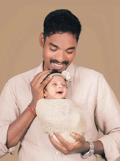 The pure joy on a new father's face as he holds his smiling, swaddled newborn daughter. A simple, heartfelt portrait against a neutral studio background.