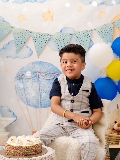 A bright smile from the four-year-old birthday boy, sitting happily in front of his cake and a backdrop of blue balloons.