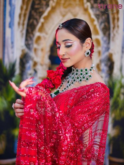 A beautiful Khaliji-inspired bridal look in a vibrant red saree. The makeup features a dramatic winged eyeliner and is paired with a stunning emerald and diamond necklace.