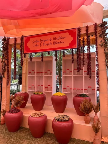 A full view of the 'Sadar Bazaar' spice stall, ready for guests to engage with. The setup includes large red urns and hanging chili strings under a draped canopy.