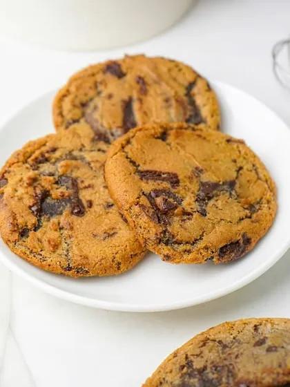 Three classic chocolate chip cookies on a white plate, with a whisk in the background to suggest they are freshly baked. This is a timeless and appealing shot for any bakery.