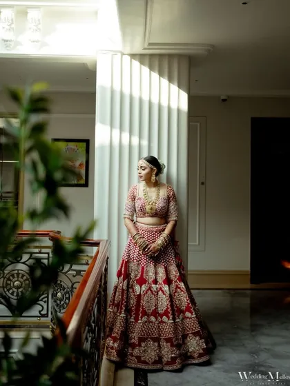 A full-length bridal portrait with beautiful natural light from a large window. The bride looks stunning in her red lehenga, framed by the modern architecture of the space.