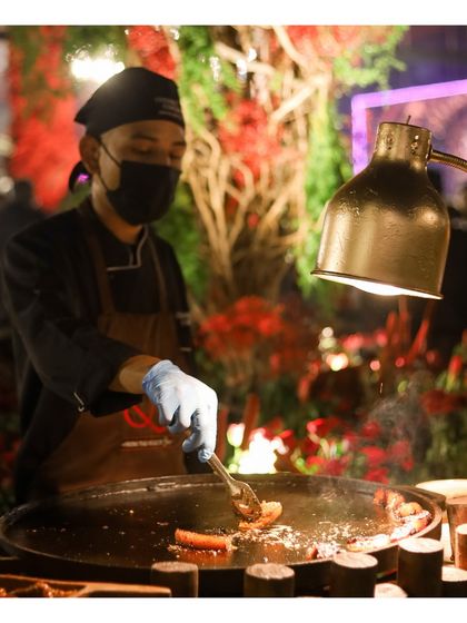 A chef at a live tawa station, illuminated by a heat lamp against a backdrop of vibrant red florals. This image captures the ambiance and professionalism of our outdoor catering setups, where every detail is considered.