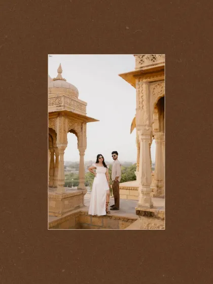 A full-length portrait of Prince and Khushi amidst the beautiful cenotaphs of Jaisalmer. The composition uses the architectural pillars to frame the couple, creating a sense of depth and grandeur.