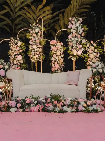 A dreamy reception stage setup in hues of pink and white. We used elegant gold arches adorned with beautiful floral arrangements and a classic white sofa to create a romantic and sophisticated photo corner for the couple.