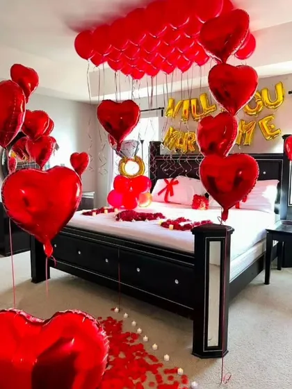 This is a classic "Will You Marry Me?" room decoration. The space is filled with red heart-shaped foil balloons, rose petals on the floor, and a bed decorated for the special moment.