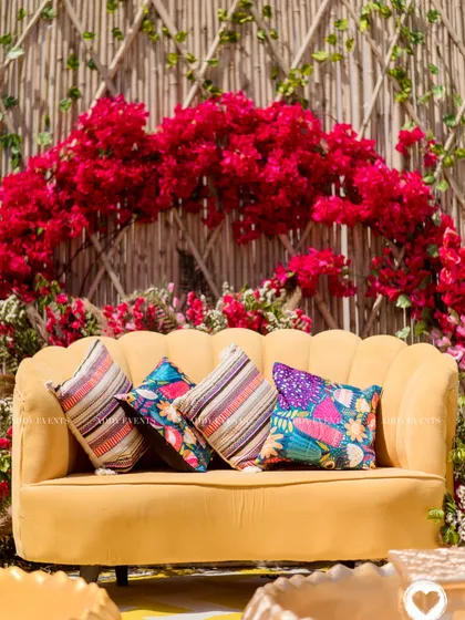 A close-up of the vibrant Haldi seating, where a mustard yellow sofa is set against a dramatic arch of red bougainvillea and a bamboo lattice wall.