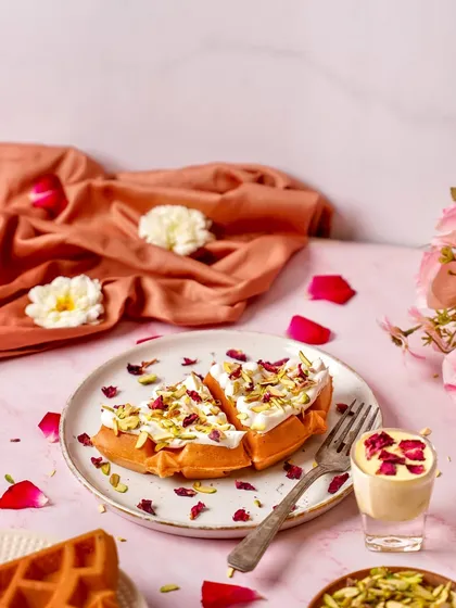 A beautiful waffle topped with cream and rose petals, shot against a soft pink background. The styling creates a romantic and delicate feel, perfect for a special menu item.