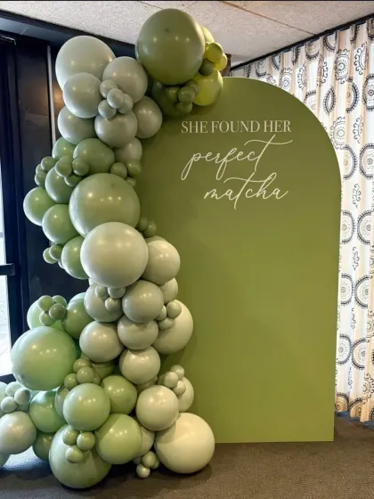 A unique and charming backdrop for a bridal shower or bachelorette party with the theme "She Found Her Perfect Matcha". The setup features a custom-shaped green backdrop and a beautiful balloon garland in shades of green.