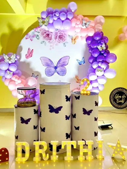 A vibrant butterfly-themed birthday for Drithi. The decor uses a purple and pink color scheme with a circular backdrop, butterfly decals on the plinths, and marquee letters for a bright and cheerful look.