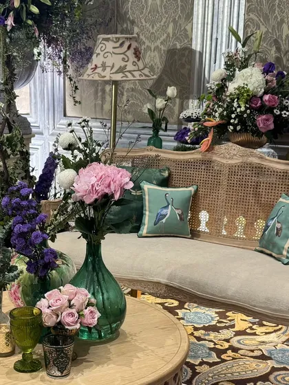 A cozy lounge corner with a cane-back sofa, bird-print cushions, and arrangements of pink and purple flowers in green glass vases.