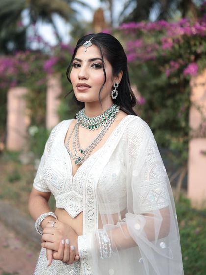 A close-up of the white lehenga with a layered diamond and emerald necklace. The outdoor setting with floral background enhances the fresh and elegant look.