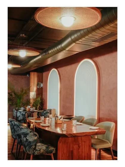 This private dining area uses arched, illuminated wall panels and floral-patterned chairs to create a more formal and exclusive feel. The exposed ductwork and rattan lights maintain a connection to the bar's overall industrial-boho aesthetic.