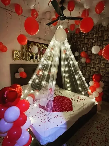A romantic birthday surprise featuring a bed decorated with rose petals, a fairy light canopy, and a room filled with red and white balloons.