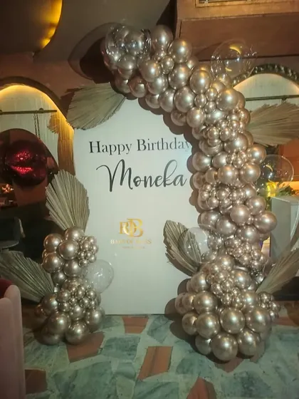 A chic and minimalist birthday setup featuring a personalized backdrop and a half-arch of metallic rose gold balloons. Dried palm leaves add a trendy, bohemian touch to the arrangement.