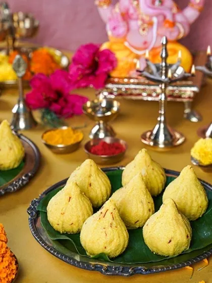 A plate of traditional, handmade yellow modaks offered to a Ganesha idol. The scene is styled with fresh flowers and brass lamps, evoking the authentic spirit of a home puja.