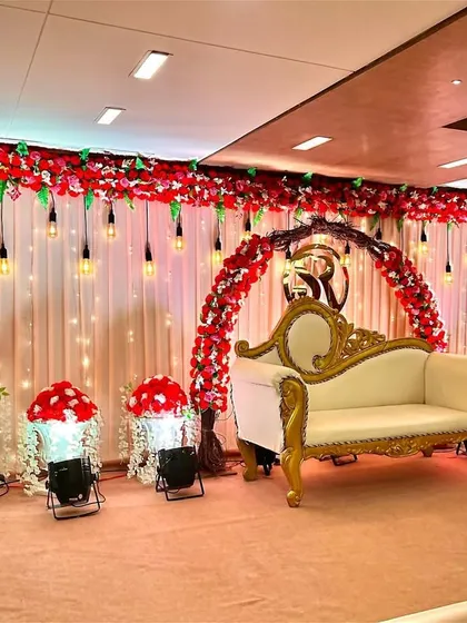 A classic and elegant stage setup inside a banquet hall. We used a backdrop of cream curtains with hanging industrial bulbs, a red floral arch, and a vintage-style sofa to create a grand and traditional look.