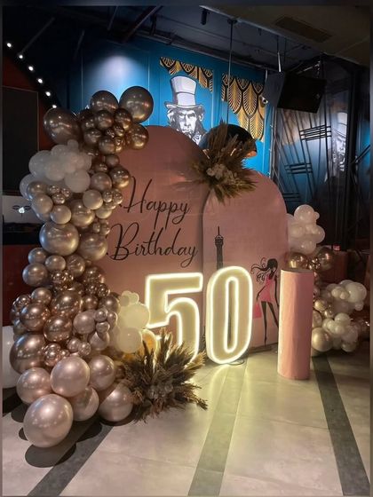 A stylish 50th birthday setup with a rose gold and blush pink theme. The decor includes a custom backdrop, neon marquee numbers, and a lush balloon garland accented with pampas grass.