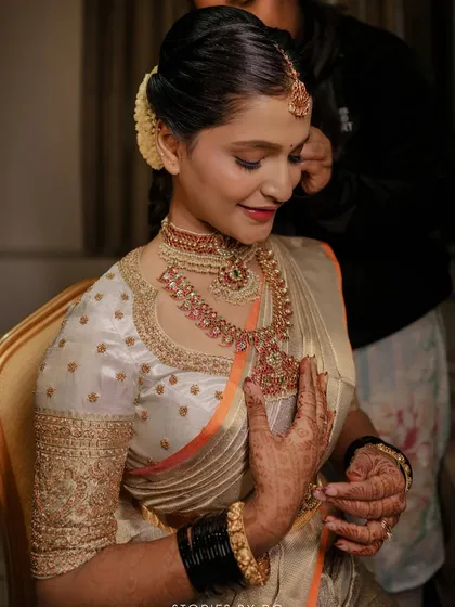 A bride getting her finishing touches, wearing a custom off-white blouse with gold embroidery. The design perfectly matches her Kanchipuram saree with its subtle orange border.