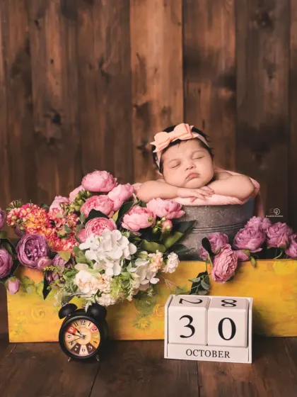 A beautiful newborn girl rests in a bucket surrounded by a bouquet of pink and purple flowers. A calendar block marks her birth date, making it a perfect birth announcement photo.