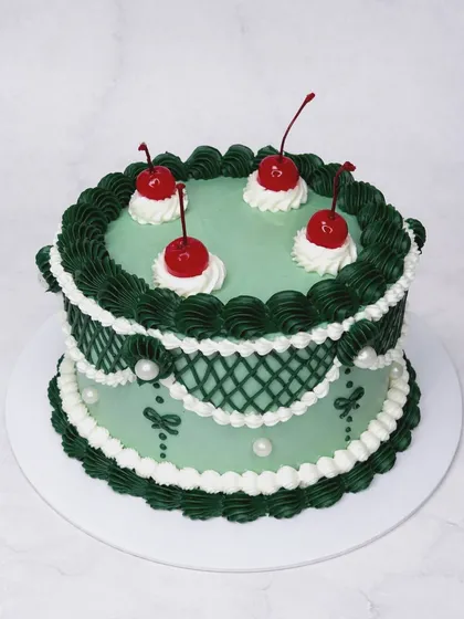 We're obsessed with the charm of vintage Lambeth cakes, and this one is a favorite. The intricate piping in forest green and white, topped with classic maraschino cherries, shows our dedication to this beautiful, ornate cake decorating style.