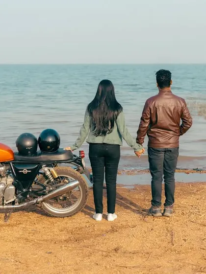 Standing hand-in-hand on a sandy shore, their Royal Enfield beside them. This image tells a story of a journey that led them to this beautiful, peaceful moment by the water.