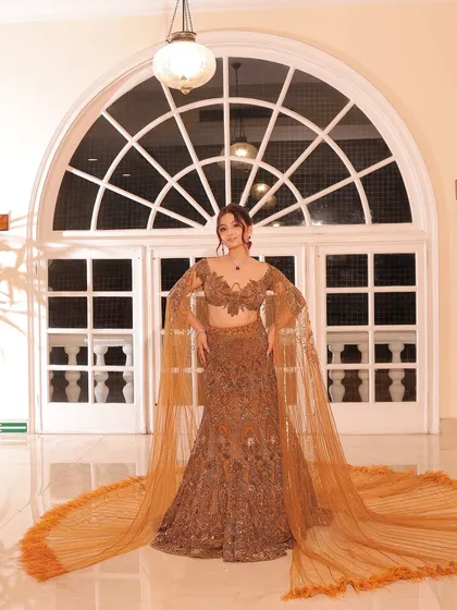 A dramatic, full-length shot of a bride in a golden-brown lehenga with a long, flowing cape.
