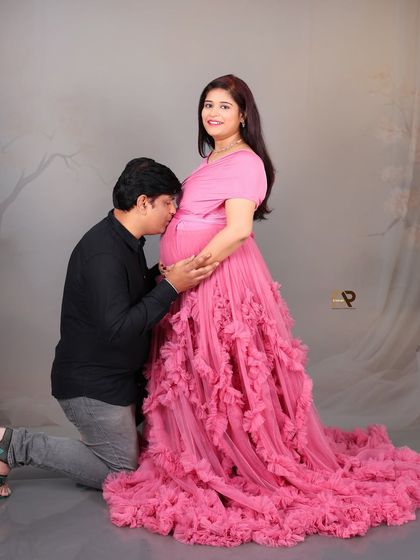 This image captures a tender moment, with the father-to-be kissing the baby bump. The vibrant pink ruffled gown adds a touch of glamour and celebration to the portrait.
