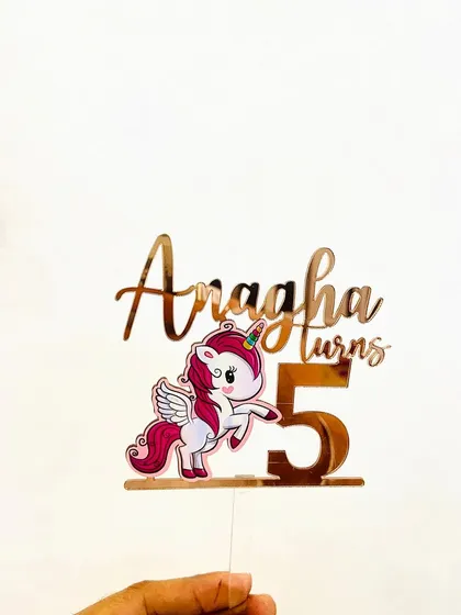 The full "Anagha turns 5" unicorn topper, held to display its complete design and size.