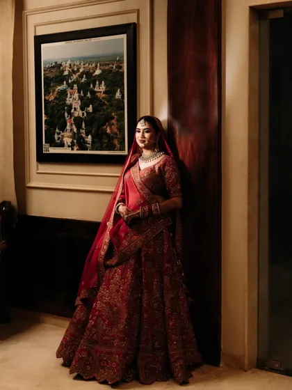 A full-length portrait of the bride in her red wedding lehenga, her poise and elegance captured in a quiet moment.