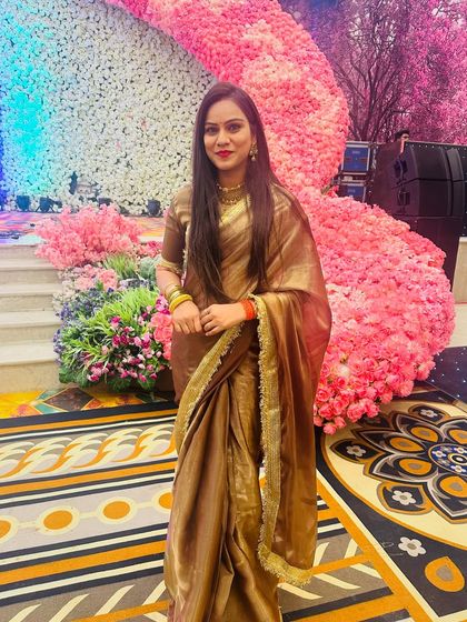 A client looking elegant at a wedding in a shimmering brown and gold tissue saree, set against a beautiful floral backdrop.